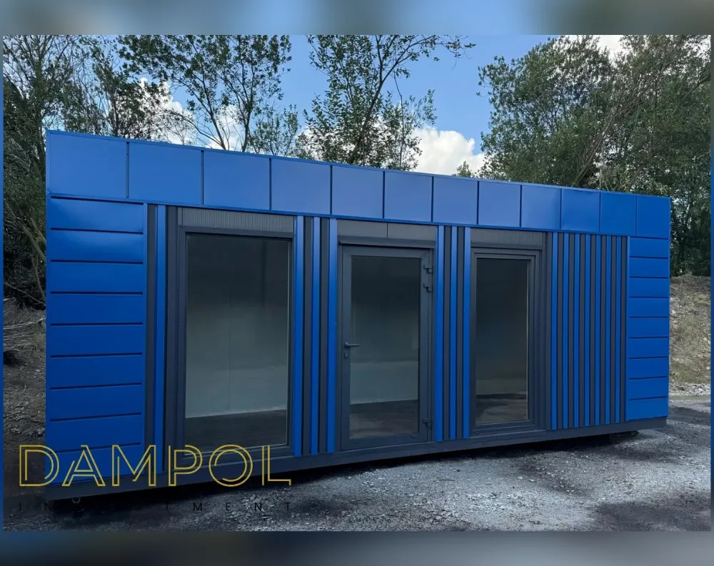 This modular building is a long, rectangular garden office clad in vivid cobalt-blue horizontal panels, with a flat, minimalist roofline finished in matching blue trim segmented into square panels. The front façade features two large fixed windows flanking a central glazed door, all framed in dark grey. Each window and the door has slim vertical anthracite pilasters between them, adding structure and rhythm to the design. The right end of the building is accented with a series of narrow vertical dark grey slats, creating a decorative textured strip that contrasts with the smooth blue paneling elsewhere. The structure sits on a low plinth, appears single-story, and has a clean, boxy, elongated shape overall.