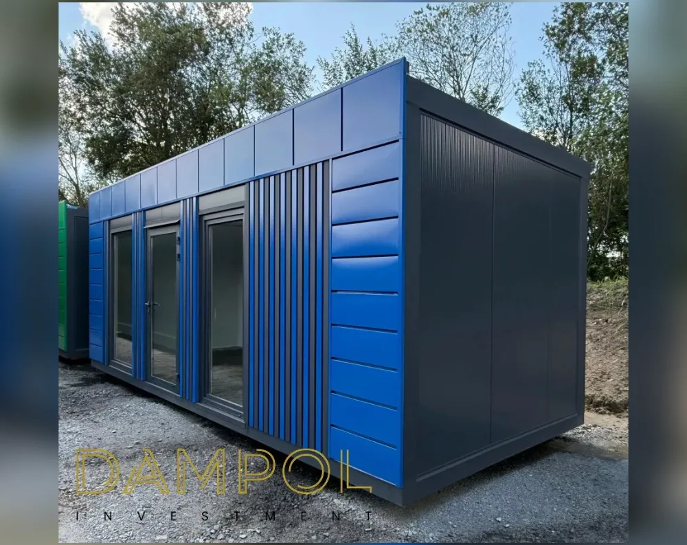 This modular garden office is a rectangular, single-story unit with a flat roof, photographed at an angle that shows both its front facade and long side wall. The front features a bold two-tone design: vivid blue horizontal paneling on the upper portion, paired with a striking vertical ribbed section running down the corner in dark charcoal. Two glass-paned doors sit side by side on the left portion of the front wall, each set within slim dark frames, allowing a view into the empty interior space. The long side wall is finished in matching dark grey corrugated-style cladding, unbroken by windows. The structure sits on gravel ground, with leafy green trees forming a natural backdrop.