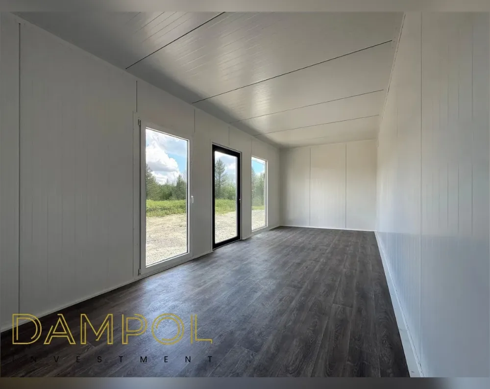This is the empty interior of a modular garden building, captured from the back corner looking toward the entrance wall. The space is long and narrow, with a slanted ceiling that slopes downward from the far end toward the entry, both finished in matching white paneling that runs along the walls as well. Dark, wood-effect laminate flooring stretches the length of the room. On the left wall near the front, there's a tall fixed window, a black-framed glass entry door, and another narrow window beside it, all offering views of gravel ground, greenery, and trees outside. The opposite wall is solid, unbroken paneling.