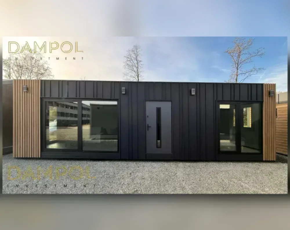 This modular building is a long, low, rectangular structure with a flat roof, presenting a sleek and contemporary profile. Its exterior is dominated by dark charcoal-black vertical panel cladding, bordered on each end by sections of warm, light wood-toned slatted paneling that add textural contrast. Centered on the façade is a single gray entry door with a narrow vertical glass strip and a slim handle. Flanking the door are two large windows: to the left, a wide sliding glass panel with a fixed pane beside it, and to the right, a matching but slightly smaller window arrangement. Small rectangular wall-mounted lights are positioned above each window, adding a modern touch.
