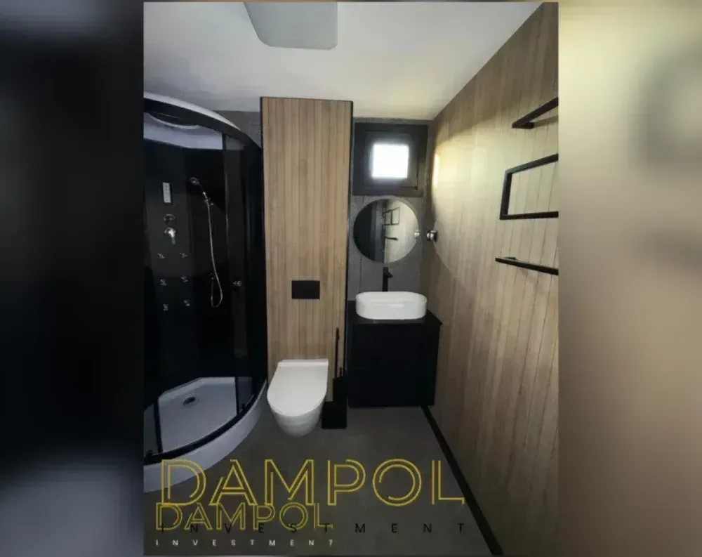 This image shows the interior of a compact bathroom rather than the exterior of a modular building. The space features a corner shower enclosure with black-framed glass panels on the left side, complete with multiple body jets and a handheld showerhead. Wood-paneled walls in a warm honey tone line the space, with one panel section housing a small recessed shelf. A round mirror hangs above a rectangular vessel sink set on a dark, floating vanity cabinet. To the left of the sink sits a modern wall-mounted toilet with a toilet brush holder beside it. A small square window is set into the wall above the vanity area, and the ceiling is white with a recessed light fixture visible.