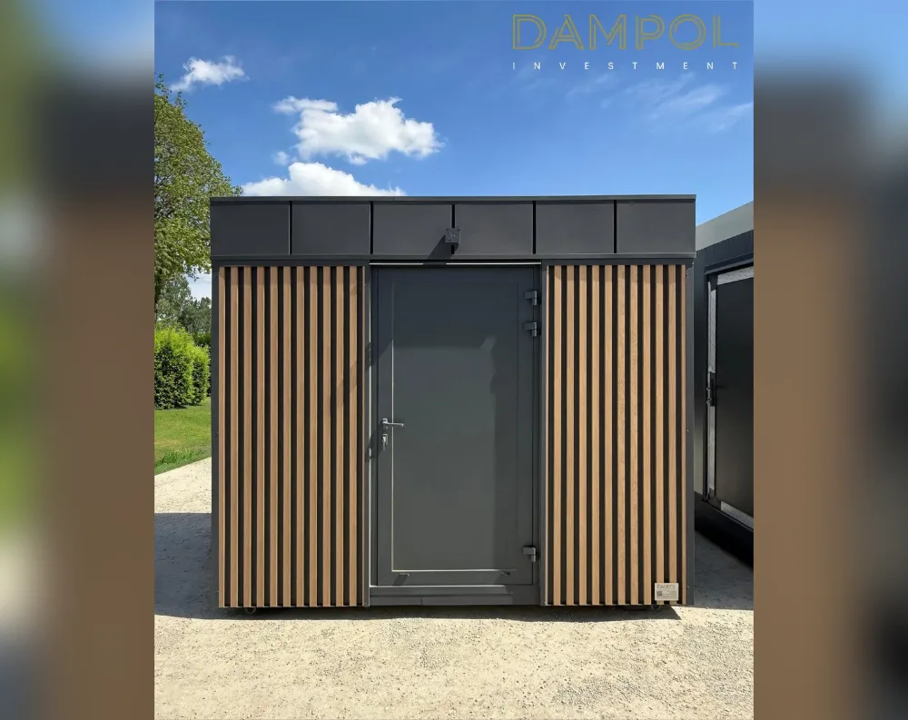 This compact modular structure has a simple rectangular shape with a flat roof, giving it a clean, boxy silhouette. The front façade features a single dark grey metal door positioned centrally, fitted with a horizontal handle and visible hinges. Flanking the door on both sides are vertical wood-toned slatted panels, alternating light and dark strips, which add warmth and texture against the otherwise charcoal-grey exterior. Above, a solid dark grey band spans the width of the building, subtly divided into panel sections. The structure sits on small wheels, suggesting portability, and is placed on a gravel surface, with greenery and a partly cloudy blue sky visible behind it.
