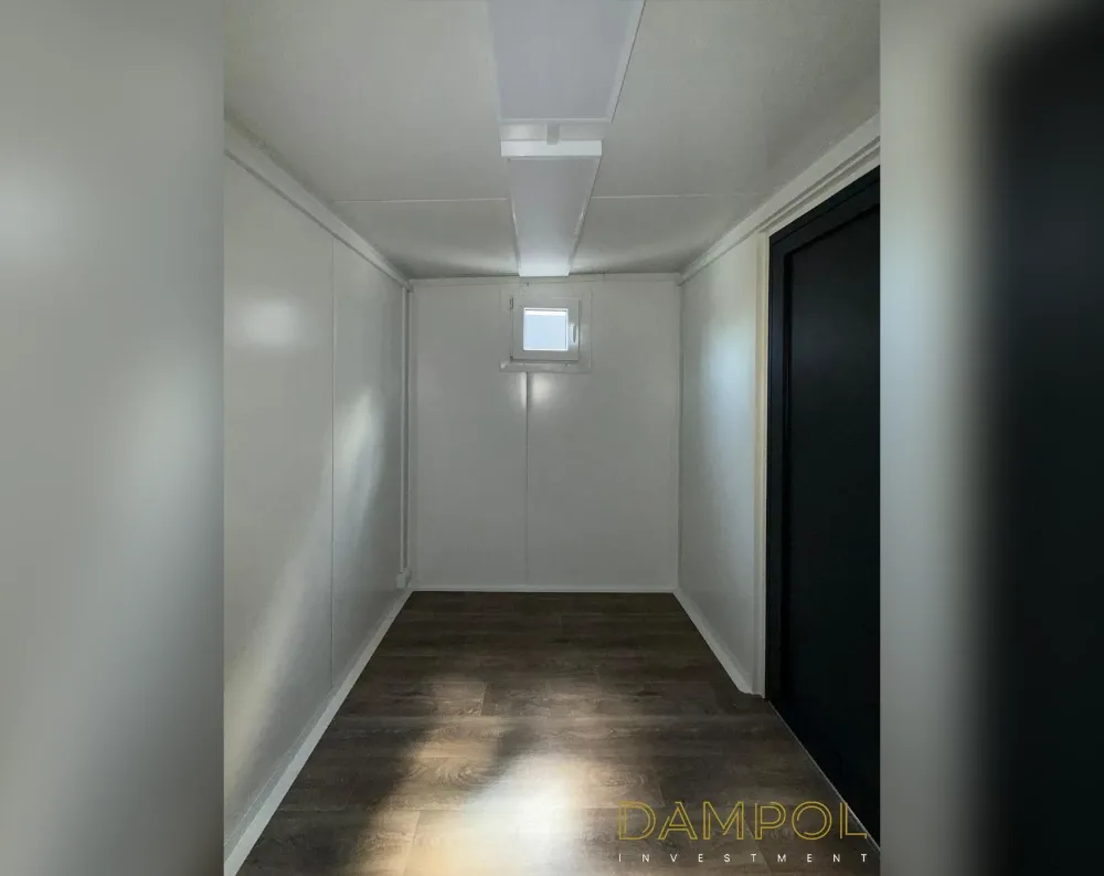 This is an interior view of a narrow modular room with a rectangular, tunnel-like proportion, longer than it is wide. The walls and ceiling are finished in smooth white paneling divided into segments, giving a clean, minimalist look. A single small square window sits centered on the far end wall, allowing natural light to spill onto the floor below. Recessed linear LED lighting runs along the ceiling panels, illuminating the space evenly. The flooring is dark brown wood-effect laminate, contrasting sharply with the pale walls. To the right, a tall dark navy or black door is set into the wall near the entrance, serving as the room's access point.