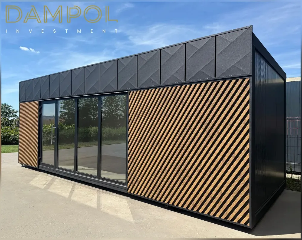 This modern modular building presents a sleek, elongated rectangular form with a flat roof, photographed at an angle that reveals both its front and side facades. The upper portion features dark gray geometric paneling with a subtle diamond-textured pattern, while the lower section combines matching dark cladding with striking diagonal wood-toned slats arranged in a chevron-like design on both the front corner and side wall. The front facade showcases a full glass frontage—a wide sliding glass door system alongside a hinged glass entry door with a vertical handle, creating an expansive, light-filled entrance. No additional windows appear on the visible side wall, which remains solid black paneling, emphasizing clean, minimalist lines throughout.