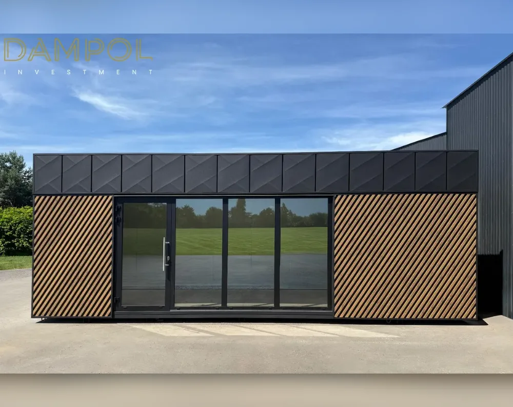 This modular building is a sleek, elongated rectangular structure with a low, flat roofline, presenting a modern, minimalist profile. The upper third of the facade features dark charcoal paneling with a distinctive diagonal diamond-textured pattern, while the lower section combines matching dark panels with warm wood-toned slats arranged in diagonal stripes on either side of the entrance. Centered on the front facade is a full-height glass entry system: a single door on the left with a metal handle, paired with three large fixed glass panels extending to the right, all framed in black. This expansive glazing reflects the surrounding grassy landscape, creating a bright, open, contemporary appearance against the black-and-wood exterior.