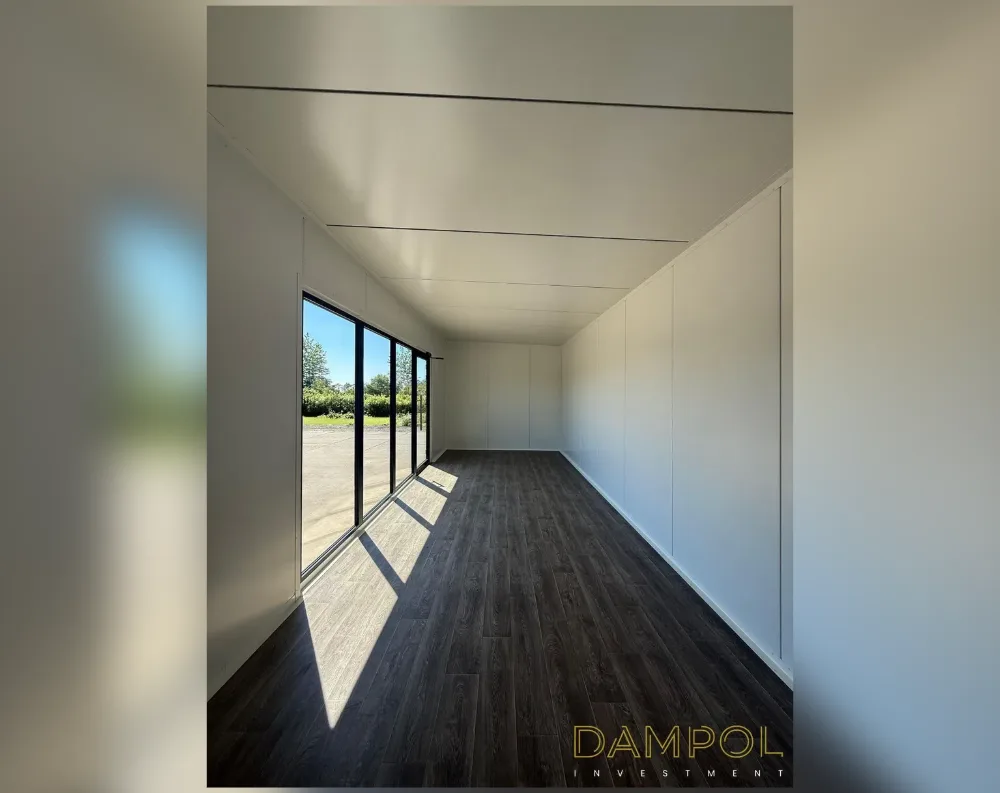 This is an interior view of a long, narrow modular room with a striking corridor-like proportion, stretching from the camera position deep into the space. The ceiling and side walls are finished in smooth, matte white paneling, divided by subtle seams that run horizontally across the ceiling and vertically down the right-hand wall, suggesting modular panel construction. The flooring is dark walnut-toned laminate with a wood-grain finish, running the length of the room and catching bright sunlight near the front. At the far left end, a full-height glazed door and window unit with black metal framing lets in natural light, revealing a paved outdoor area, trees, and blue sky beyond.