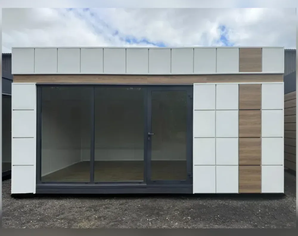 This modular garden office presents a clean, rectangular silhouette with a flat roof and boxy proportions, wider than it is tall. The exterior cladding combines white paneled squares with warm wood-toned vertical accents, creating a modern grid pattern across the upper band and side sections. The front facade is dominated by a large glass expanse: a wide sliding glass door system spans the left and center, paired with a single hinged glass door with a metal handle on the right side of the glazed area. Dark grey aluminum framing outlines all glass elements, contrasting sharply with the light exterior panels. Solid white-and-wood paneled sections flank both sides of the glazed front, adding structure and visual balance.