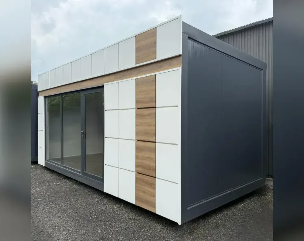 This modular garden office is a rectangular, single-story structure with a flat, slightly sloped roof that extends slightly beyond the walls. The front façade features a full-width glass section, including a double sliding glass door flanked by a fixed glass panel, allowing clear visibility inside. Above this glazing runs a band of narrow horizontal white panels, interrupted by a vertical strip of wood-toned paneling. To the right of the entrance, alternating vertical panels of white and light oak-effect finish create a distinctive geometric accent. The building's side and rear are clad in smooth dark charcoal-grey paneling, giving a sleek, modern contrast to the patterned front.