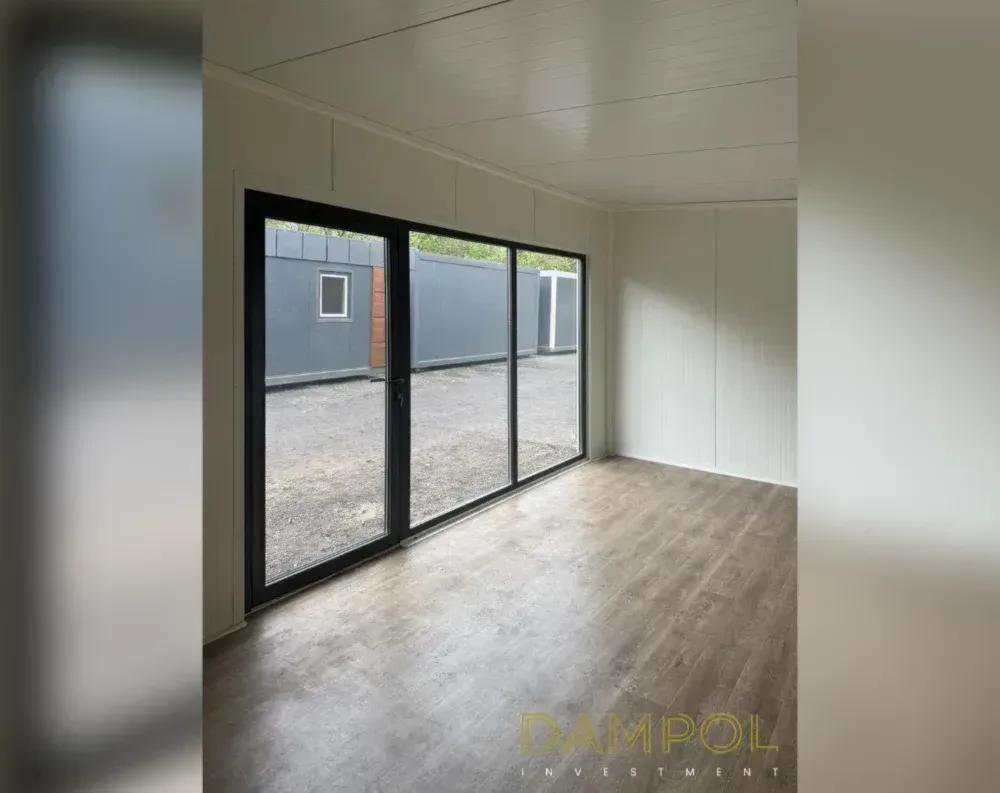 This image shows the interior of a modular garden office, an empty rectangular room with a clean, minimalist finish. The walls and ceiling are clad in smooth cream-white paneling with visible horizontal seams, giving a tidy, uniform appearance. A wood-effect vinyl floor in warm grey-brown tones runs throughout, adding warmth to the space. On the left wall stands a large black-framed glass door system, comprising a hinged door and adjoining fixed glass panels, allowing daylight to flood in and offering a view of an adjacent grey-paneled structure outside. The room appears bright, open, and unfurnished, emphasizing its versatile, ready-to-use layout.