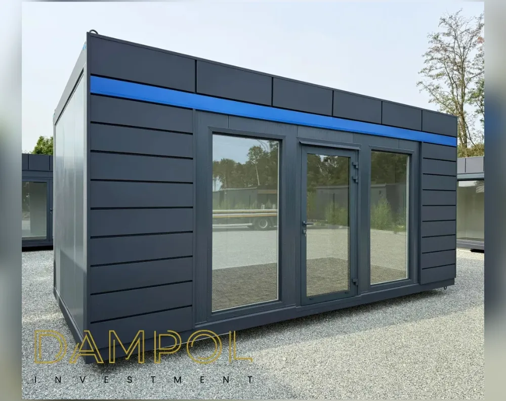 This modular building has a sleek, rectangular shape with a low-profile, flat roof, giving it a modern, minimalist silhouette. The exterior is clad in horizontal dark charcoal-grey paneling, creating a sharp, structured texture across the walls. A vivid blue horizontal stripe runs along the upper edge of the front and side facades, just beneath the roofline, adding a bold accent to the otherwise monochrome design. The front features a large glass door in the center, flanked by two expansive fixed glass window panels, all framed in matching dark grey. This wide glazing invites natural light and offers a clear view of the reflected trees and surroundings outside.
