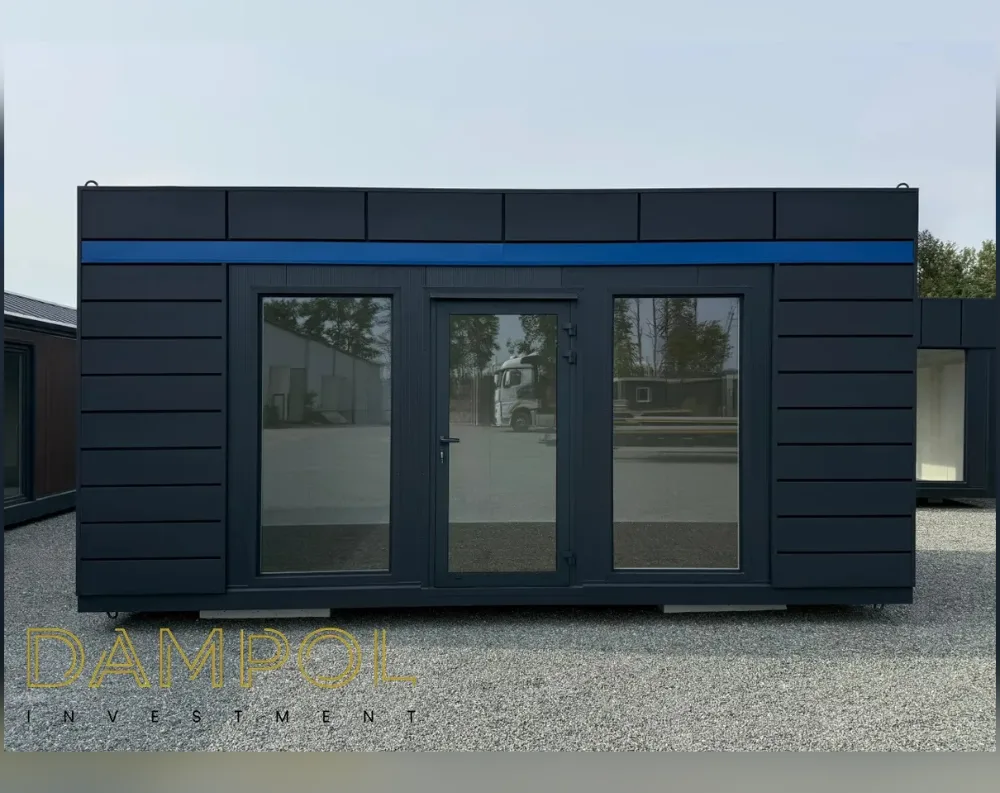 This modular building has a long, low, rectangular shape with a flat roof, giving it a sleek, contemporary profile. Its exterior is clad in dark charcoal, horizontally grooved paneling that runs the full length of the structure, broken by a bold blue horizontal stripe just below the roofline for a striking accent. The front façade features a symmetrical layout: a large fixed glass window on the left, a central glazed door with a slim black frame, and a matching large window on the right, all sharing the same dark tinted glass. The unit rests on visible support blocks, and small lifting rings are fixed at the top corners of the roof.