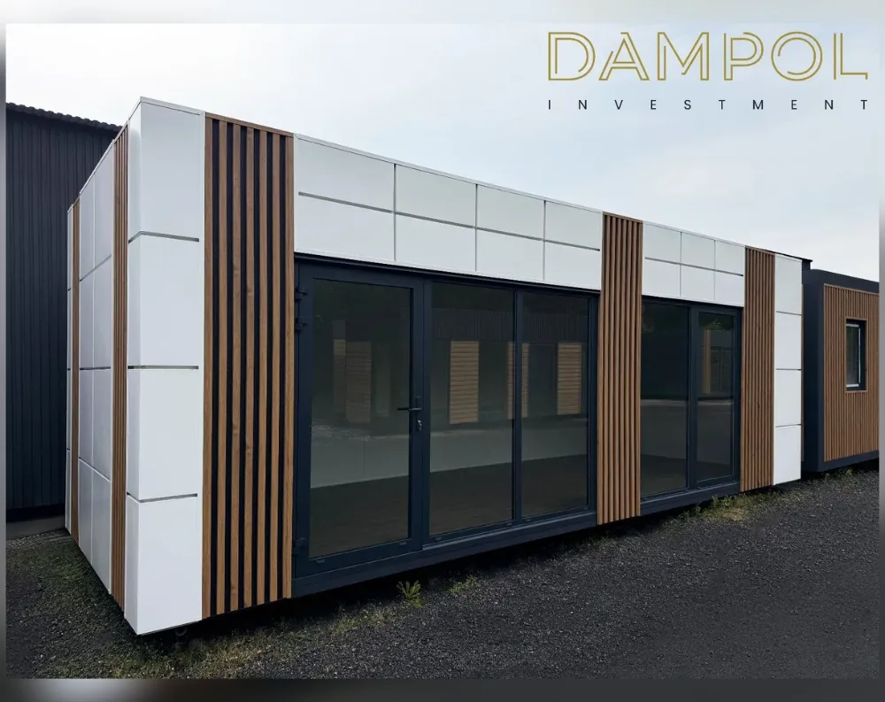 This modular building is a long, low, rectangular structure with a flat roof, photographed at an angle that shows its front and side. The exterior combines smooth white panel cladding with vertical strips of natural wood-toned slats, arranged in alternating bands for a striking contrast. The front facade features a large glass door system—paired dark-framed doors flanked by wide glass panels—stretching almost the full width, allowing generous light into the interior. Above this glazing runs a row of square white paneling. A small additional window appears on the adjoining side structure. The overall proportions are elongated and single-story, giving it a sleek, modern cabin-like appearance, sitting on a gravel surface with a plain corrugated shed visible in the background.