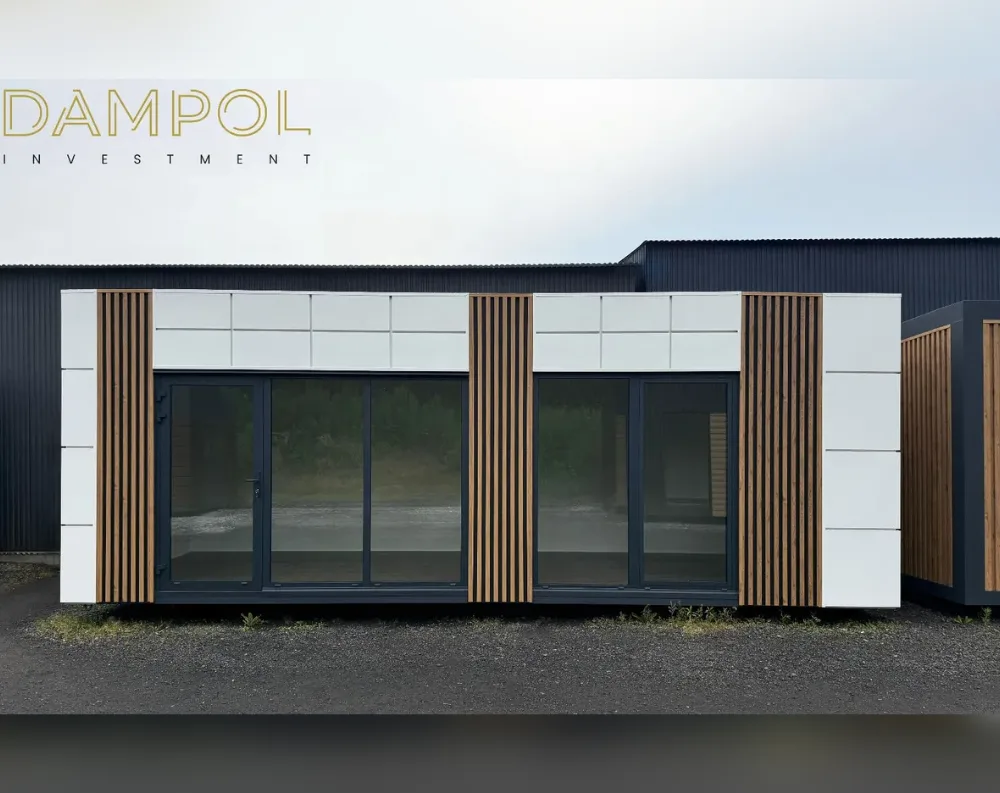 This modular building presents a long, low rectangular form with a flat roof, sitting on gravel ground. Its façade alternates between smooth white panels and vertical wood-slat cladding, creating rhythmic sections that divide the structure visually. Two large glass sliding door units dominate the front, each flanked by dark-framed panels, with a single hinged door visible on the left side leading inside. The upper portion features a grid of square white panels running the length of the building, contrasting with the darker window frames below. Warm timber battens frame both ends and the central divide, adding texture against the crisp white surfaces, giving the structure a modern, minimalist aesthetic with natural accents.