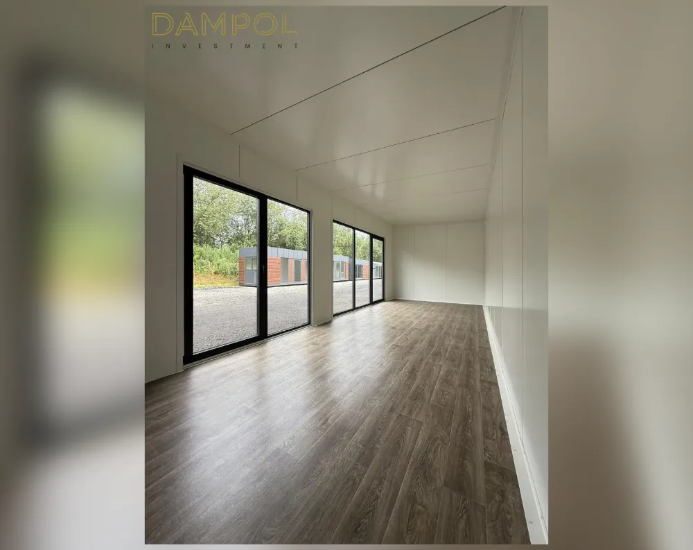 This photo shows the bright, empty interior of a long, narrow modular room with a slightly angled ceiling that slopes upward toward the back, creating a subtle vaulted effect. The walls and ceiling are finished in smooth white paneling, while the floor features warm, dark wood-look laminate planks running the length of the space. Two sets of tall, black-framed glazed sliding doors sit along the left wall, each divided into two panels, allowing natural light to flood in and offering a view of a gravel lot and another modular building outside. The far end of the room narrows into a bright, open corridor-like extension, giving the space a clean, minimalist, and airy feel.
