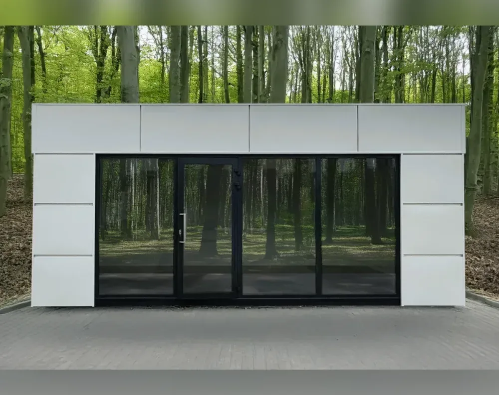 This modular garden building sits low and wide, a single-story rectangular structure with a flat roofline. Its cladding is smooth white paneling, divided into clean geometric sections, with the upper portion forming a broad fascia band that overhangs slightly above the glazed front. The front facade is dominated by black-framed glass: a central hinged door flanked by two large fixed or sliding glass panels on either side, all trimmed in matching black aluminum. Narrow white paneled sections bookend the glass on both the left and right, each featuring subtle horizontal reveal lines for texture. The structure stands on a paved area, surrounded by a dense backdrop of tall, slender forest trees.