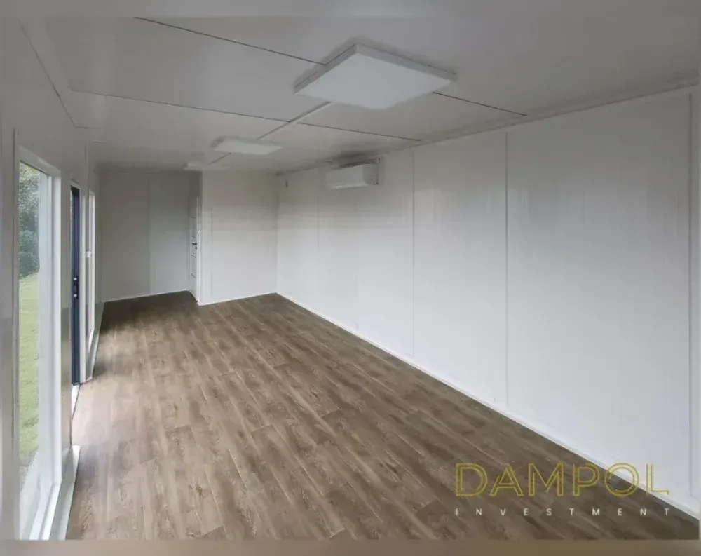 This image shows the bright, empty interior of a modular building rather than its exterior. The elongated rectangular room features warm brown wood-effect laminate flooring contrasting with crisp white wall panels and a matching white paneled ceiling divided into sections. Natural light floods in through a tall glass door and adjacent window on the left side, offering a view of green grass outside. Overhead, two square recessed light fixtures illuminate the space, while a wall-mounted split-system air conditioning unit sits high on the right wall. Toward the back, a doorway leads to an adjoining room. The space appears clean, minimalist, and ready for customization, with simple functional fittings and no additional furniture or decor visible.