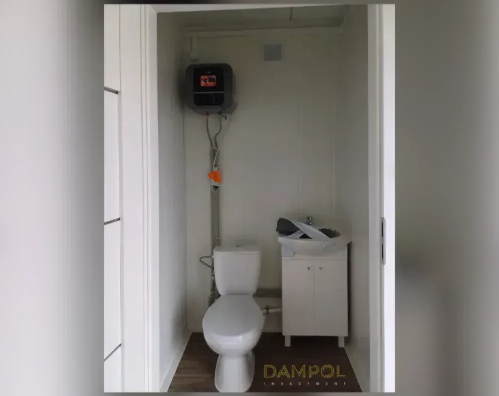 This photo shows the interior of a compact modular bathroom rather than the building's exterior. The narrow, tall space features light beige paneled walls and ceiling with visible seams, giving it a simple cabin-like finish. A small square ventilation grille sits high on the back wall. Below it, a wall-mounted electric water heater with a digital display feeds pipework running down to a white toilet positioned against the rear wall. To the right stands a compact white vanity cabinet with double doors, topped by a sink still wrapped in protective packaging. The floor is dark wood-toned laminate, and the doorway framing is bright white, creating clean, minimal proportions throughout.