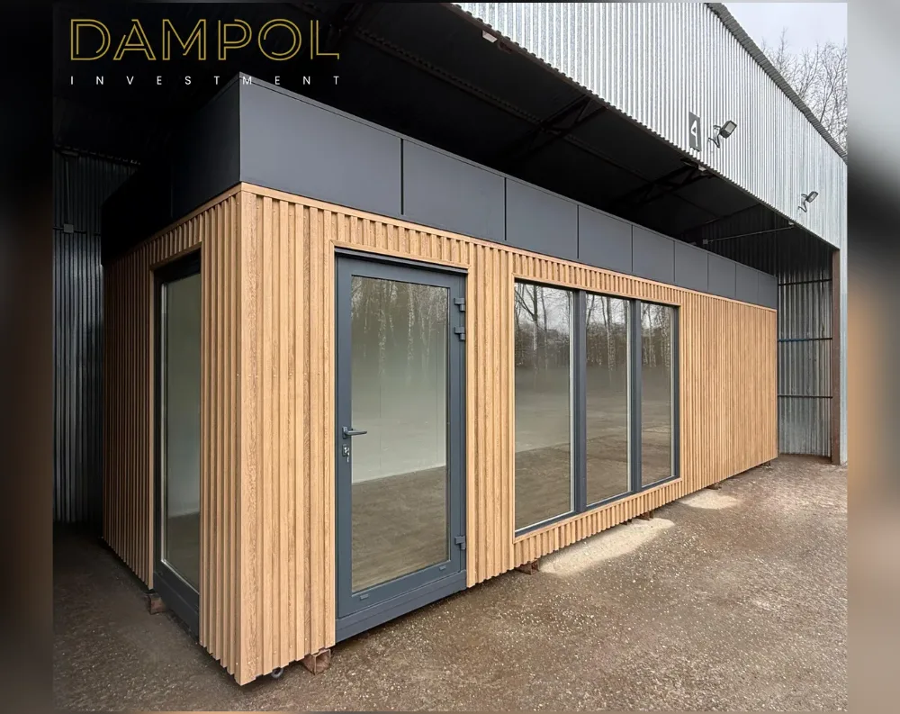 This modular building is a sleek, rectangular structure with a low-profile flat roof and a dark charcoal fascia band running along its top edge. The exterior is clad in vertical light oak wood slats, giving it a warm, contemporary look that contrasts with the black upper trim. The front-facing wall features a full-height glass entry door with slim dark metal framing, positioned left-of-center, flanked by a tall fixed window to its left and two wide floor-to-ceiling glass panels to the right, offering an unobstructed view inside. The structure sits on raised metal footings, and its clean lines and minimalist palette give it a modern, cabin-like presence against the industrial warehouse backdrop.