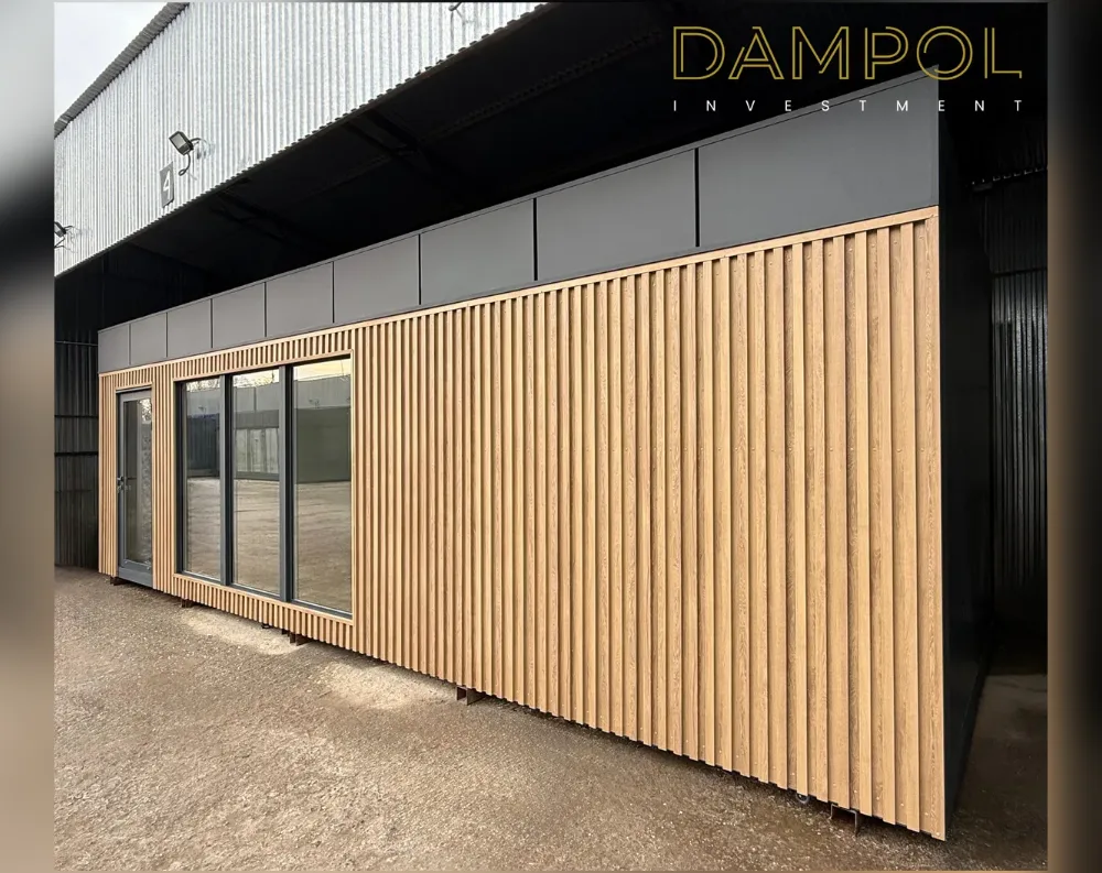 This modular garden office is a long, low rectangular structure with clean, contemporary lines. Its facade is dominated by vertical wooden slat cladding in a warm honey-oak tone, running floor to roofline in narrow evenly-spaced battens. A dark charcoal-grey band caps the upper portion of the wall and wraps the flat roofline, creating strong contrast with the wood below. On the left side sits a fully glazed door with slim black framing, beside three large glass window panels that let in ample light. The structure stands on visible metal support feet, elevating it slightly off the gravel ground, giving it a sleek, floating appearance.