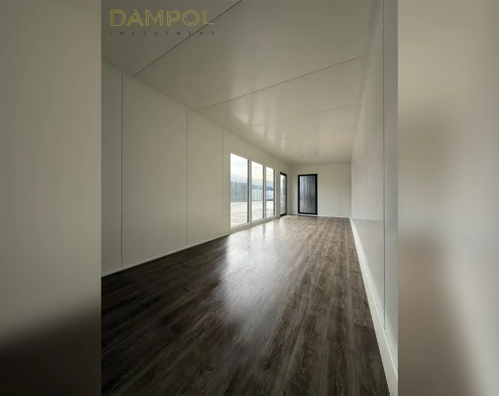 This photo shows the interior of a long, narrow modular room with a rectangular footprint and a flat ceiling that gives it a clean, minimalist feel. The walls and ceiling are finished in smooth white paneling, divided into simple rectangular sections. The floor is a rich dark wood-look laminate, with a glossy sheen reflecting the light. On the left-hand wall near the far end are three tall vertical windows set close together, allowing daylight to spill in and reveal a hint of an outdoor structure beyond. Just past the windows sits a single dark-framed glass door, matching the modern, understated aesthetic of the space.