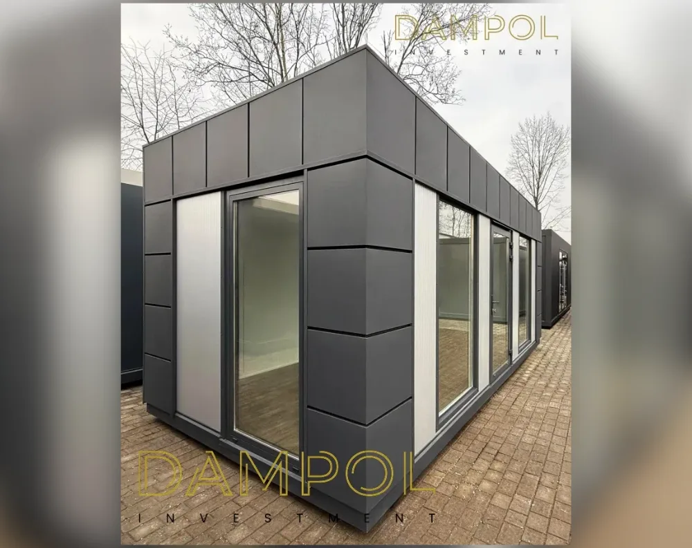 This modern modular structure sits as a long rectangular box with a flat roof, photographed at an angle showing both its short front end and long side elevation. The cladding is dark charcoal-grey composite panels arranged in vertical sections, giving a sleek, sharp-edged appearance with visible seam lines. Slim vertical white or light-grey accent strips run alongside the corners and glazing, adding contrast. The front facade features a large floor-to-ceiling glass door flanked by narrow glass panels, while the long side wall includes a wide run of glass doors and windows—appearing to be four to five panes plus a door—allowing full visibility into the empty white-walled interior. The building rests on a paved brick surface, with bare trees visible behind it.