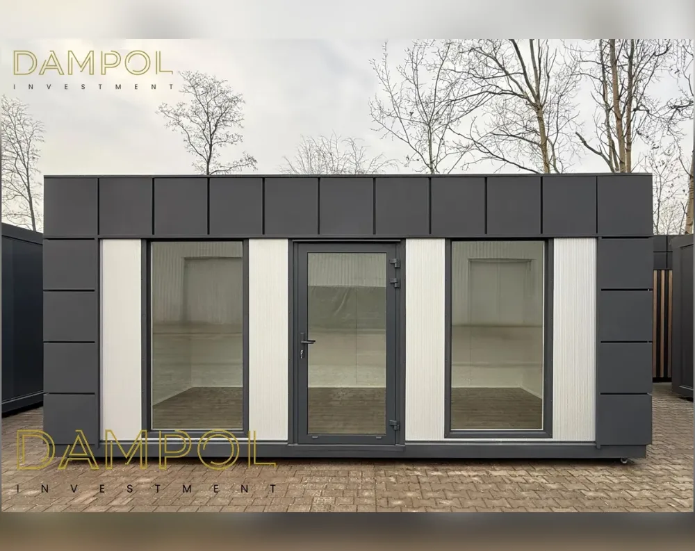 This compact rectangular modular building has a low, flat-roofed profile with a horizontal emphasis, wider than it is tall. The upper band of the facade is clad in dark charcoal-grey paneling divided into vertical segments, running the full width like a header strip. Below this, the wall alternates between matching dark grey corner panels and lighter, ridged off-white vertical panels. Centered on the front is a dark-framed glass entry door with a slim handle, flanked symmetrically by two large fixed picture windows, each bordered by white paneling. The overall look is sleek, minimalist, and modern, sitting on small visible wheels, with a paved brick surface beneath and bare trees in the background.