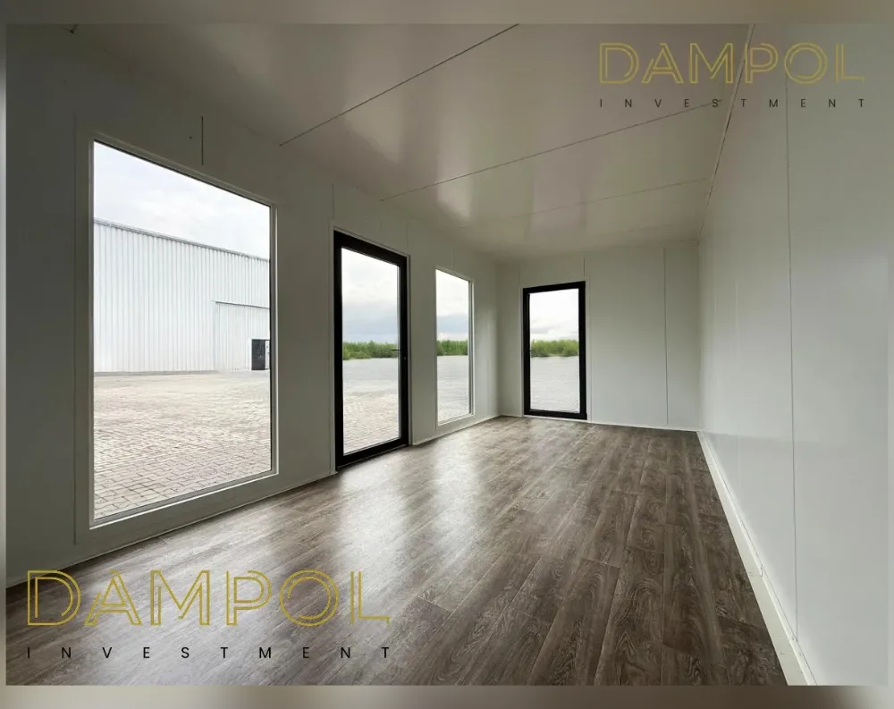 This is an interior view of an empty modular room with a clean, minimalist finish. The walls and ceiling are smooth white panels, and the floor is laid with rich, dark brown wood-effect laminate running diagonally across the space. Along the left wall, a large fixed window offers a view of a warehouse building and paved yard. Further along, a tall glazed door with black framing sits beside another full-height window, both showing an open outdoor area with greenery. At the far end, a narrower black-framed window lets in additional light. The room has a rectangular, elongated shape with high ceilings, giving it an airy, bright, and spacious feel throughout.