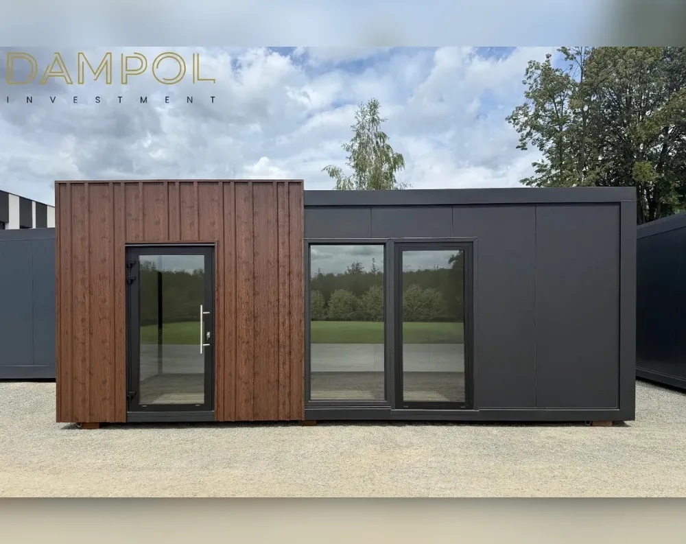 This modular garden building is a long, low rectangular structure with a flat roof, standing on gravel ground with trees and greenery behind it. On the left, a section is clad in vertical dark wood-toned paneling, featuring a tall glass-paneled door with a slim black frame and metal handle. The right two-thirds of the building are covered in smooth charcoal-grey panels, broken by two large windows: one fixed wide pane and one narrower door-style window, both framed in black. The overall proportions are wide and horizontal, with a slightly overhanging flat roofline, giving it a sleek, modern, minimalist appearance with contrasting wood and dark grey finishes.
