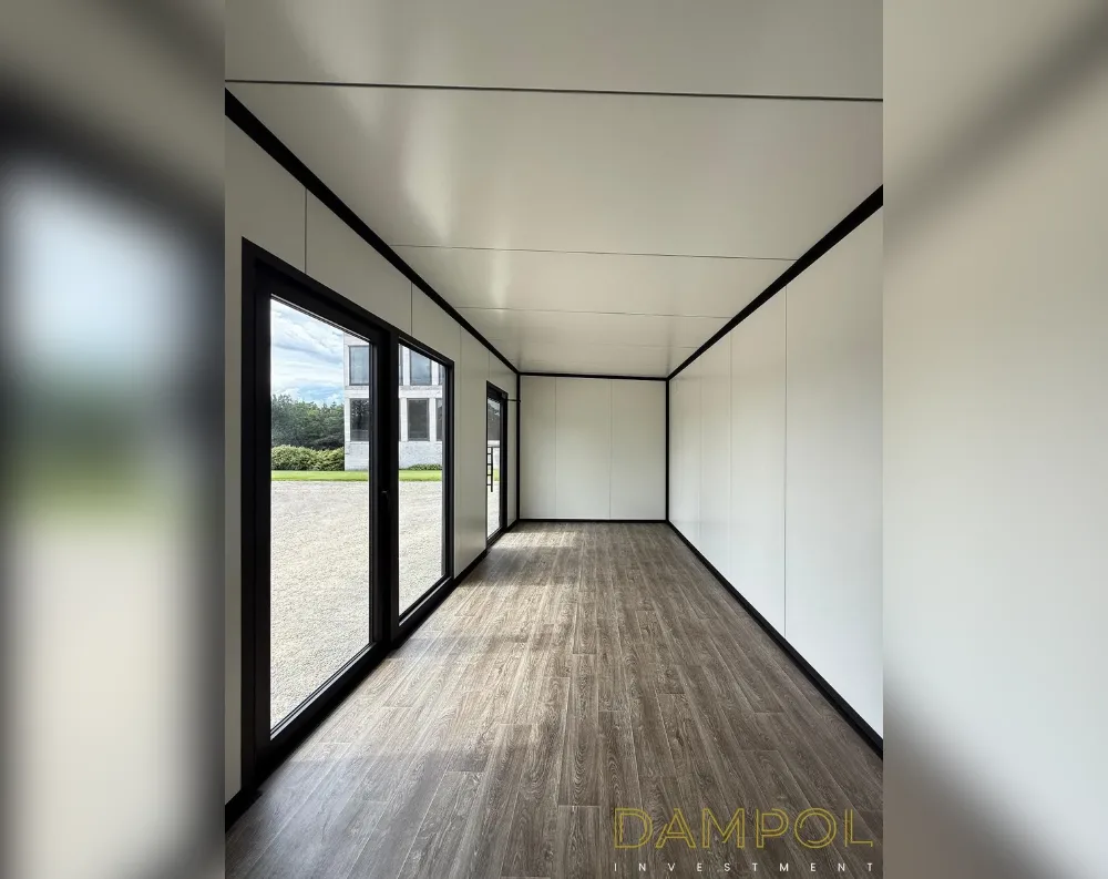 This photo shows the empty interior of a long, narrow modular building, likely a garden office or portable unit, viewed from one end down its length. The space has a rectangular footprint with a flat white ceiling and matching white wall panels divided by black trim seams that run along the corners and joints. The flooring is a warm grey-brown wood-effect laminate, laid in linear planks that draw the eye toward the far wall. On the left side, a full-height glass door with black framing opens near the entrance, followed by a second glass panel further along, allowing daylight and a view of gravel ground and greenery outside. The far end wall is solid white, unbroken by windows.