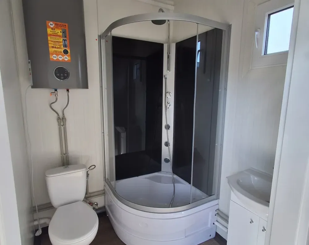 This compact bathroom occupies a corner of the modular building, dominated by a curved corner shower enclosure with dark smoky glass panels and a silver aluminum frame, set into a white basin tub. Above it, a chrome showerhead extends from an arched top rail, with a handheld sprayer and control dials mounted on the shower's interior post. To the left, a compact wall-mounted water heater with a control panel sits above exposed plumbing pipes leading down to a white toilet. On the right, a small white pedestal-style sink with cabinet storage beneath sits near a small window with a black frame, allowing natural light into the space. Walls are finished in pale cream paneling throughout.