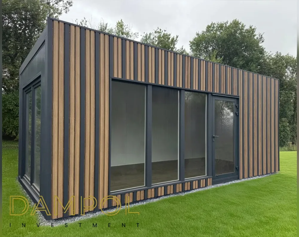 This modular garden building sits on a lush green lawn, its rectangular form topped with a flat, slightly sloped roof that gives it a low, contemporary profile. The exterior combines dark charcoal-grey paneling with vertical slats of natural wood-toned cladding, creating a striking striped effect across every wall. The front façade features three large fixed glass panels and a single glazed door with dark metal framing, all set within a wide floor-to-ceiling window arrangement. The angled corner reveals additional narrow window panels wrapping around the side. A thin gravel border lines the base, separating the structure from the surrounding grass, while tall trees form a green backdrop behind the building.