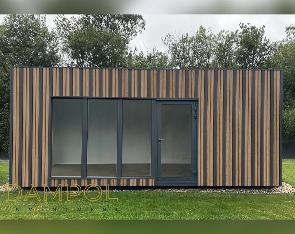 This modern garden office is a low, rectangular structure with a flat roof, wider than it is tall, giving it a sleek, horizontal profile. Its exterior is clad in vertical wooden slats alternating with dark charcoal-grey panels, creating a striped, contemporary look across the entire facade. On the front wall, three large fixed glass window panels sit side by side on the left, filling the space with light, while a single dark-framed glazed door with a slim handle is positioned to the right, matching the grey trim. The structure rests on a bed of pale gravel, surrounded by green lawn, with dense trees forming a leafy backdrop under an overcast sky.