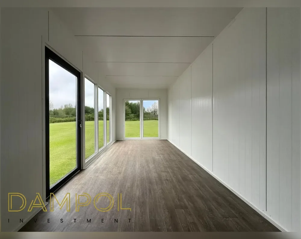 This is an interior view of a long, narrow rectangular modular room with a low, flat ceiling and clean white paneled walls running its full length. On the left side, a row of floor-to-ceiling windows and a full-height glass door with dark frames line the wall, overlooking a bright green lawn and distant trees. At the far end, centered, is another set of double glass doors with white frames, also opening onto grass and greenery, flooding the space with natural light. The floor is finished in dark walnut-toned wood-effect planking, contrasting with the bright white walls and ceiling. The overall look is minimalist, airy, and unfurnished, emphasizing the room's depth and connection to the outdoors.