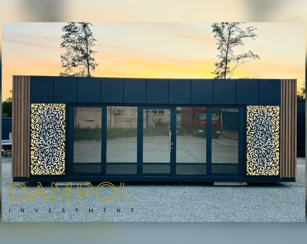 This modern modular garden building has a low, elongated rectangular shape with a flat roof, wider than it is tall. The facade is finished in dark navy-blue paneling, framed on each side by vertical wood-slat cladding that adds warmth and texture. Dominating the front is a wide glass sliding door and window system divided into five panels, with a central hinged door featuring a slim vertical handle, all set in matching dark frames. Flanking the glass are two decorative panels with intricate cut-out leaf and vine patterns, backlit with a warm golden glow. The structure sits on a raised base, with a soft sunset sky and silhouetted trees visible in the background.