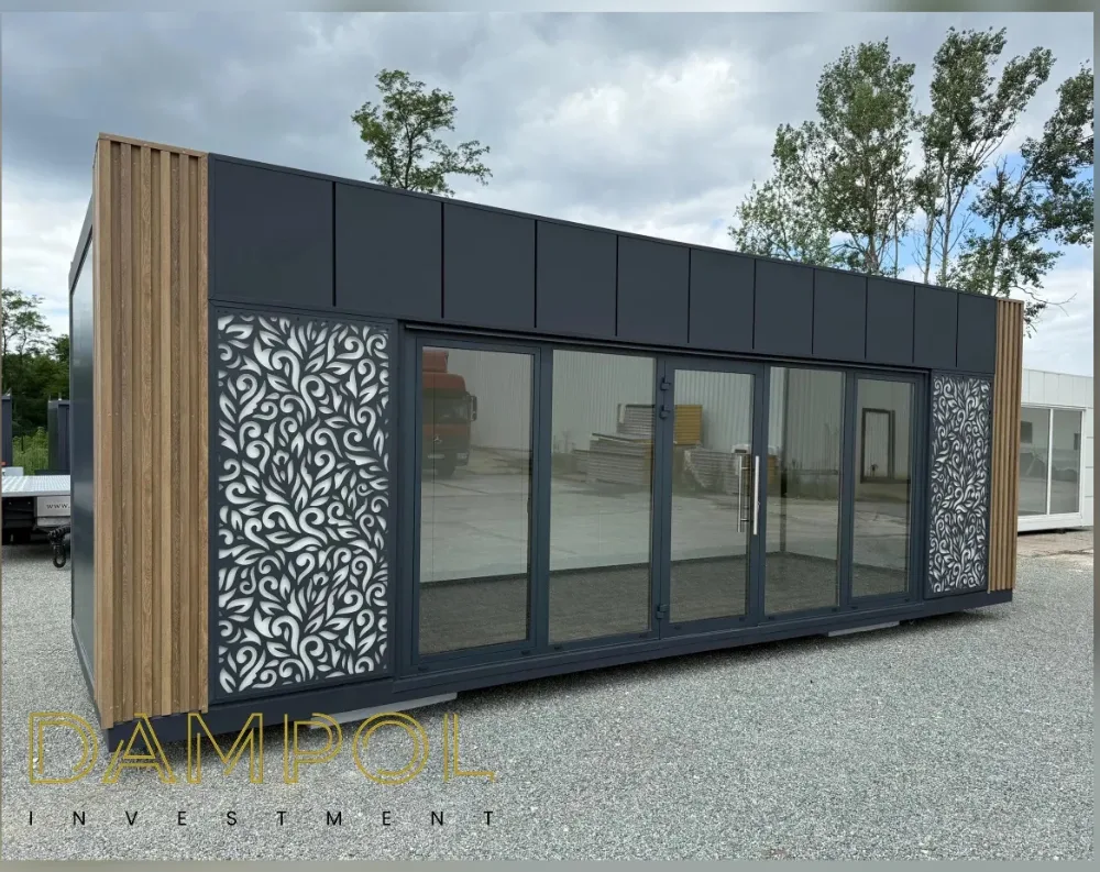 This modular garden office is a sleek, elongated rectangular structure with a low-profile flat roof, photographed at an angle that reveals its full length. The exterior cladding is dark charcoal-grey panelling, contrasted by vertical wooden slat accents on both the near corner and far end. The front façade features a wide expanse of glazing: four large fixed glass panels flank a central double glass door, creating a bright, open entrance. Two decorative panels with intricate white floral or vine-like laser-cut patterns are mounted on dark backgrounds, positioned symmetrically on either side of the glass frontage. The structure sits on a paved gravel lot, with trees and additional containers visible in the background.