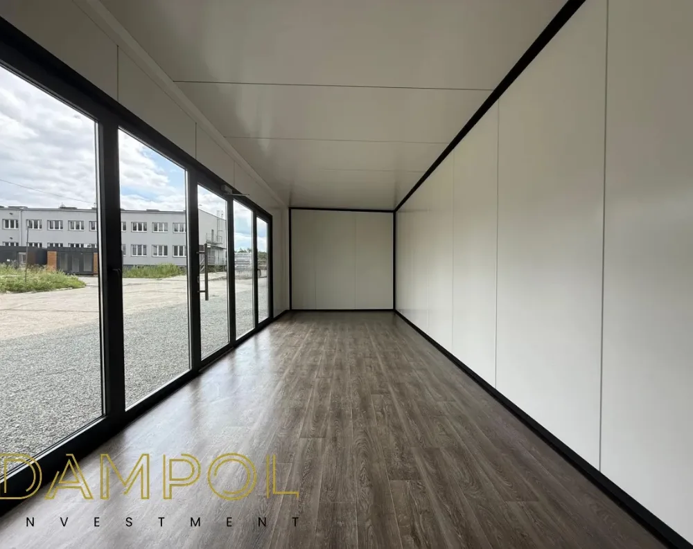 This photo shows the interior of a long, narrow modular building, viewed down its length toward a closed end wall. The left side features a full run of floor-to-ceiling glass panels with dark, slim frames, including a sliding glass door, allowing abundant natural light and a view of a gravel yard and a neighboring building outside. The right side has solid, smooth white wall paneling extending the full length. The ceiling is flat and white, divided by subtle seams, with a black trim strip running along the top edge where it meets the wall. The flooring is dark brown wood-look laminate, and the far end wall is a plain white partition with a black-framed edge.
