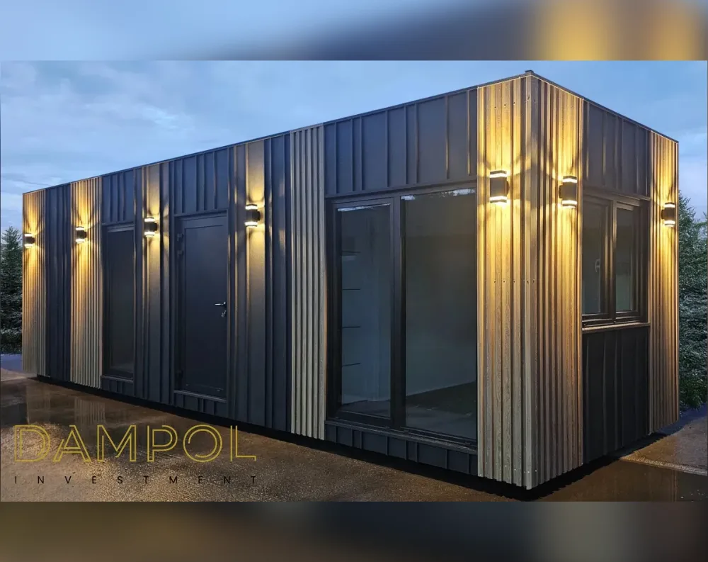 This modular building is a long, narrow rectangular structure with a flat, single-slope roof, photographed at dusk. Its exterior is finished in dark charcoal vertical corrugated cladding, contrasted by warm wood-toned vertical slat paneling at both corners and around select openings. Wall-mounted lantern-style light fixtures line the facade, casting soft upward and downward glows on the wood paneling. The front-facing side shows two dark doors and two narrow windows, while the long side features a large sliding glass door and an adjacent expansive window, both framed in black. The structure sits on a paved surface, with evergreen trees visible in the background against a soft blue evening sky.