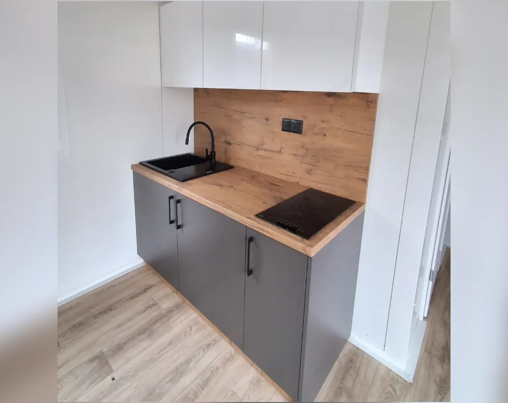 This photo shows a compact kitchenette rather than an exterior building view, so I'll describe what's actually visible. The space features a small corner kitchen unit with glossy white upper cabinets mounted against a white wall, paired with matte dark grey/charcoal lower cabinets featuring sleek black bar handles. A warm wood-grain countertop and matching backsplash panel run along the wall, housing a black rectangular sink with a curved black faucet and a black induction cooktop. A light wood-toned laminate floor extends across the room, and a wall-mounted electrical outlet sits above the counter. The walls are plain white, giving the space a clean, minimalist, modern aesthetic.