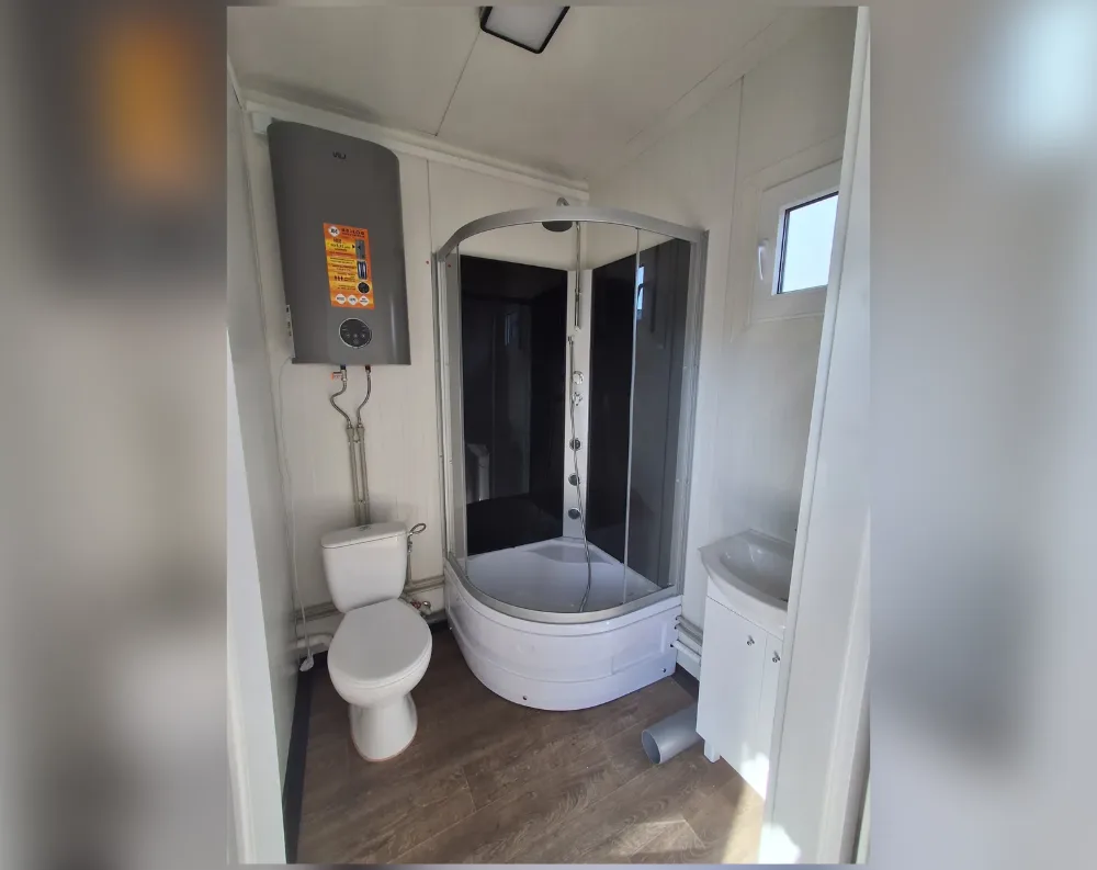 This compact modular bathroom unit features a narrow, tall rectangular layout with light beige/cream cladded walls and ceiling panels. A single small rectangular window sits high on the right wall, letting in daylight, while the entry doorway is implied at the image's foreground. The space is dominated by a corner-mounted shower enclosure with curved, frosted-glass sliding doors and a white basin base, positioned centrally-right. To its left stands a compact wall-mounted water heater with an orange instruction label, feeding pipes down to a white toilet below. A small white sink with cabinet storage sits to the right of the shower. Flooring is brown wood-effect laminate, and a flat ceiling panel with a recessed light fixture completes the utilitarian, space-efficient design.
