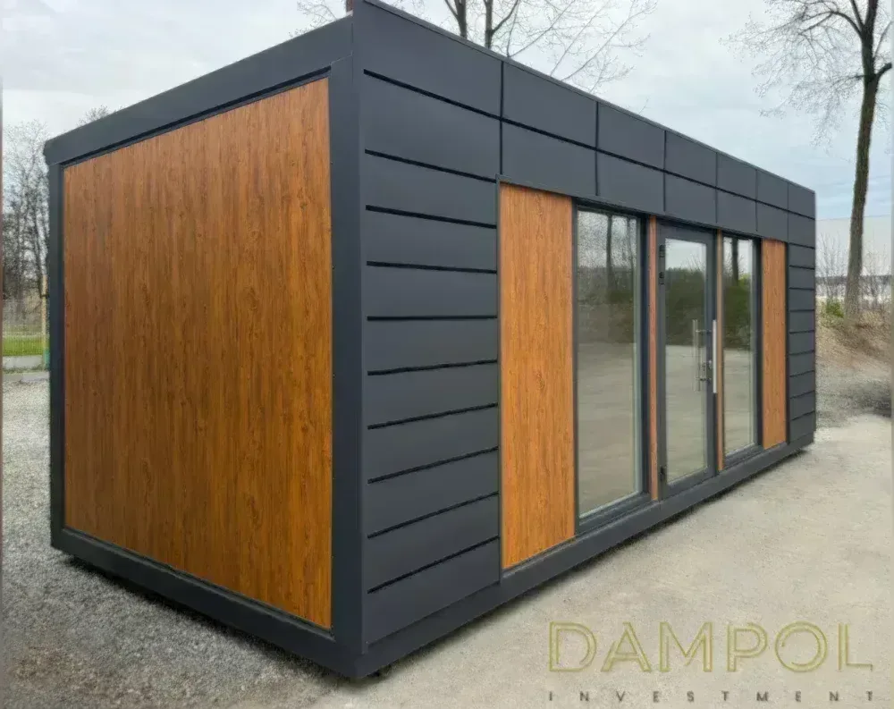 This modular garden office is a sleek, rectangular structure with a flat roof, photographed at an angle that reveals both its long side and front façade. The exterior combines dark charcoal-grey paneling, arranged in horizontal slats, with warm honey-toned wood-effect panels used as accent strips—one large panel on the side, and two flanking the entrance. The front features a full glass door with a vertical handle, positioned centrally, flanked by two large fixed glass window panels, allowing ample natural light. Above the door, a slim horizontal window strip runs along the upper façade. The building sits on gravel ground, with bare trees and a cloudy sky in the background.