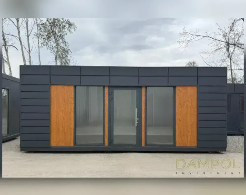 This modular building is a sleek, rectangular structure with a low-profile flat roof and a horizontal design emphasis. The exterior cladding is finished in dark charcoal grey paneling with subtle horizontal ridges, giving it a modern, streamlined look. Centered on the front façade is a glass-paneled entry door with a metal handle, flanked symmetrically by two large fixed windows offering clear views inside. On the outer edges of the window sections are vertical wood-toned accent panels, adding warmth and contrast to the otherwise cool-toned exterior. Above the windows and door, the upper portion of the wall continues in the same dark cladding, reinforcing the clean, boxy proportions of the unit.