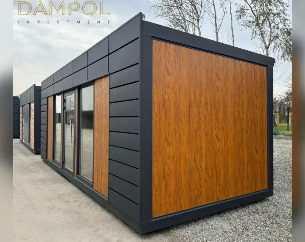 This modular building presents a rectangular, single-story structure with a low-pitched flat roof and clean, elongated proportions. The exterior combines dark charcoal horizontal paneling with warm honey-toned wood-grain accent panels, creating contrast on the front face and side wall. The front facade features a glass double door flanked by a large fixed window pane, all set within slim black frames, positioned toward the left side of the unit. A matching wood-panel strip sits beside the entrance. The long side wall showcases an expansive wood-textured panel bordered by dark trim. The building sits on a gravel lot, with a partially visible second unit behind it, echoing the same design.
