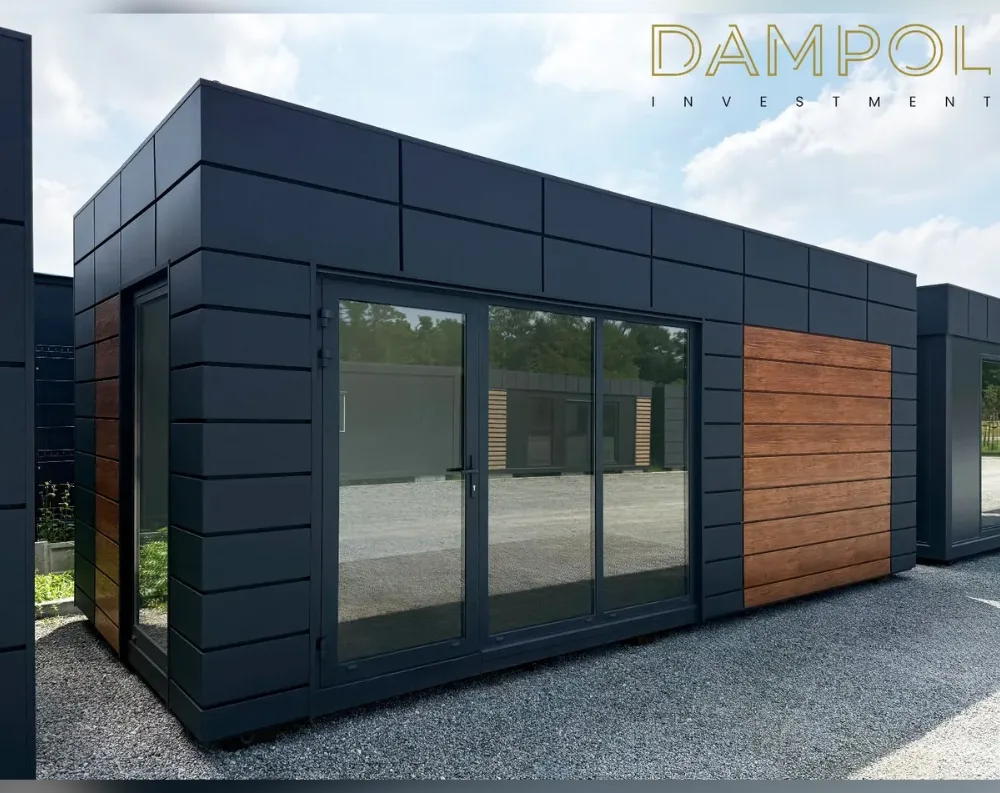 This modular building is a sleek, rectangular structure with a flat roof, clad entirely in dark charcoal-grey paneling arranged in a horizontal ribbed pattern that gives it a modern, streamlined look. The front façade features a large sliding glass door system with three panels, positioned centrally, allowing full visibility into the interior space. To its left stands a narrower single-hinged glass door, tucked into the corner of the building. Breaking up the dark exterior, a warm honey-toned wood-paneled accent section sits to the right of the sliding doors, adding contrast and texture. The structure rests on a gravel surface, with trees and additional matching units visible in the background.