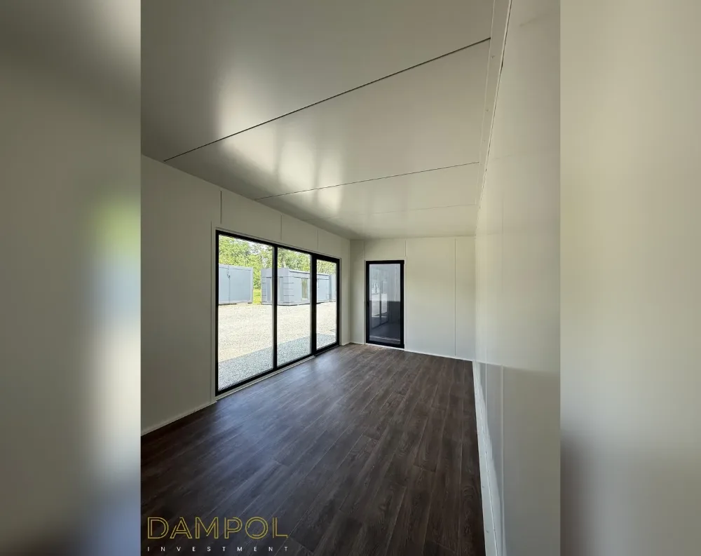 This interior view shows an empty modular room with a sloped ceiling that rises toward the back, giving the space an airy, elongated feel. Dark walnut-toned laminate flooring runs the length of the room, contrasting with the crisp white wall and ceiling panels. On the left wall, a large sliding glass door with slim black framing spans three panels, offering a view of gravel ground and other modular units outside. Beside it, a narrower black-framed window sits closer to the corner. The walls appear smooth and seamless, finished in a matte white panel material, giving the space a clean, minimalist, contemporary look typical of a modern garden office or modular building interior.