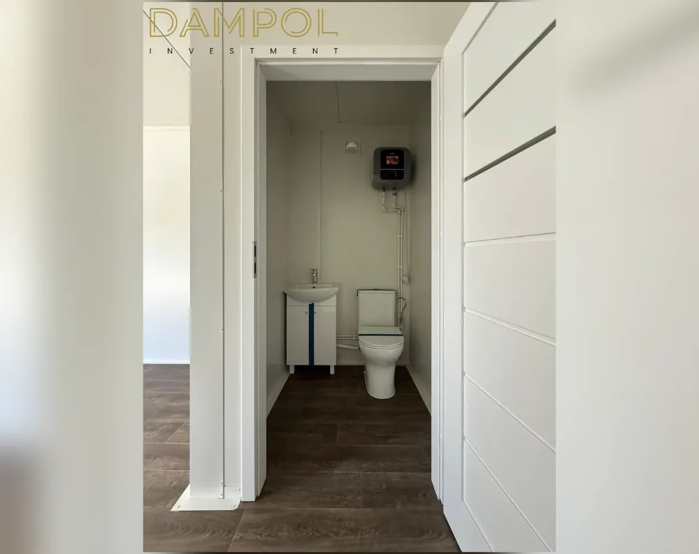 This photo shows an interior view through an open doorway into a compact bathroom within a modular structure. The white door swings open toward the viewer, revealing a narrow rectangular room with pale cream-colored wall paneling. Dark wood-effect tile flooring runs throughout the space, continuing into the adjacent room visible on the left. Inside the bathroom, a small pedestal sink with a navy-accented cabinet sits beside a standard toilet, with visible plumbing pipes running up the wall to a compact water heater unit mounted near the ceiling. A small square vent or fixture appears above. The overall proportions suggest an efficient, single-room utility space with simple, functional finishes.