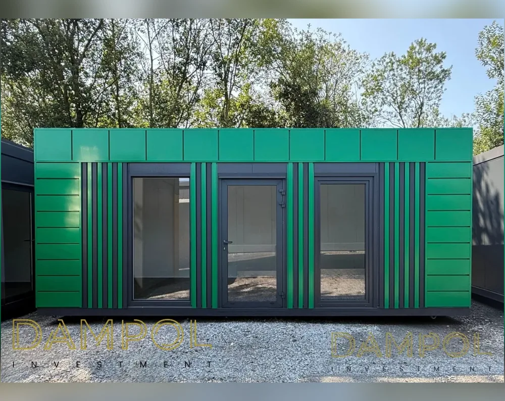 This modular garden office is a long, rectangular single-storey unit with a flat roof, standing on a gravel surface against a backdrop of leafy trees. Its cladding is vibrant green paneling on the upper section and sides, broken by vertical dark charcoal-grey stripes that add rhythmic contrast. The front façade features a symmetrical layout: two large fixed glass windows on either side flank a central glazed door, all framed in matching dark grey. The overall proportions are wide and low, emphasizing horizontal lines. Thin structural seams divide the green panels into a grid pattern, giving a clean, modern, prefabricated appearance suited for a compact office or garden studio.