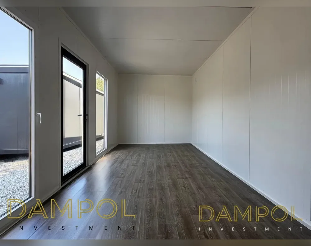 This is an interior view of a modular garden office, empty and ready for finishing. The room is long and rectangular with a low, flat ceiling made of pale paneling with visible seams. Dark wood-toned laminate flooring runs the length of the space, contrasting sharply with the crisp white wall panels that line both sides. On the left wall, a tall glass door with black framing sits beside a narrow vertical window, both offering views of a gravel yard and neighboring structure outside. Natural light floods in from this side, while the opposite wall remains solid and windowless, creating a bright, open, minimalist feel throughout the room.