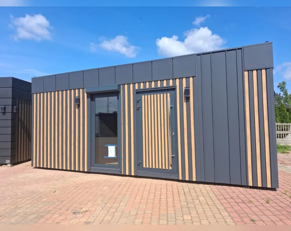 This modular garden office is a long, low rectangular unit with a flat roof, photographed at an angle that shows its full front façade and part of one side. The exterior is finished in dark charcoal-grey panelling, accented with vertical slatted wood-toned strips arranged in evenly spaced columns, particularly framing the entrance and window areas. The front features one large fixed window with slim grey framing on the left side, and a matching grey door with a small vertical window pane to its right, both flanked by wooden slats. Small rectangular wall-mounted lights sit above each opening. The building rests on a paved brick surface, with another dark unit visible to the left.