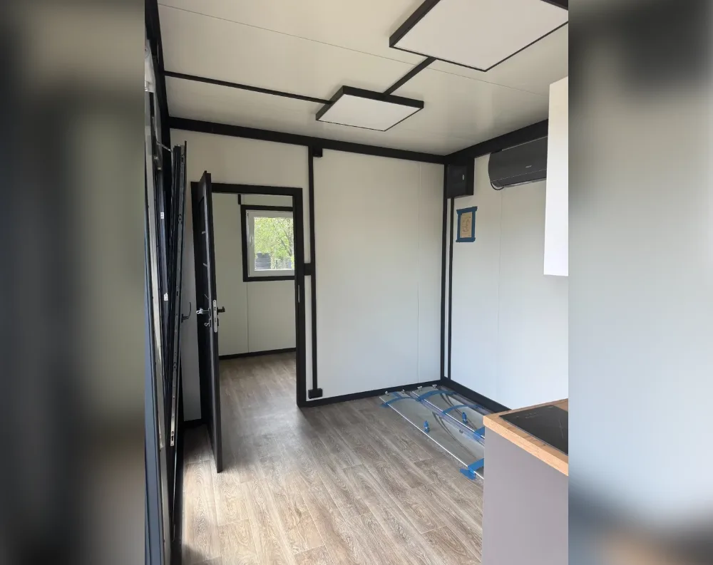 This is an interior view of a modular garden office, showing a narrow rectangular room with light wood-effect laminate flooring and off-white wall panels bordered by dark charcoal trim strips that outline the ceiling, corners, and panel seams. Two flat recessed ceiling light panels illuminate the space, alongside a wall-mounted air conditioning unit in the upper right. A single interior door with a small square window stands open on the left, leading to a corridor with an exterior window revealing greenery outside. In the foreground, a partial kitchen counter with a built-in stovetop is visible, its surface still protected with blue tape.