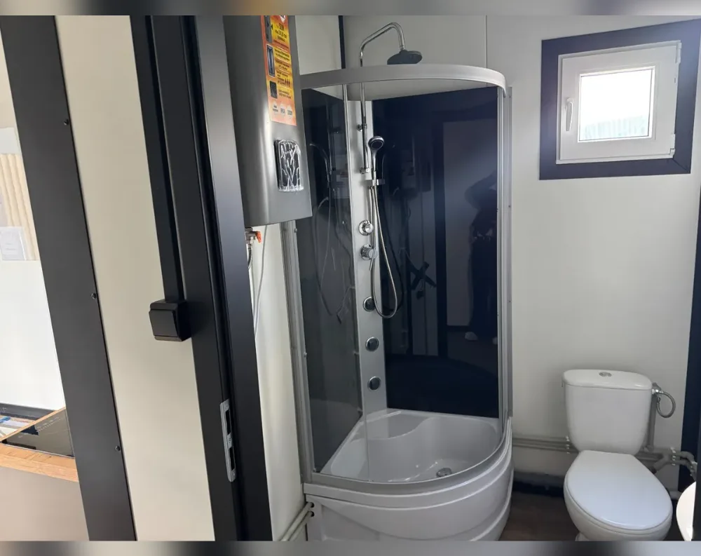 This photo shows the interior of a compact bathroom module rather than the building's exterior. A corner-mounted shower cabin with curved smoked-glass paneling dominates the space, fitted with a chrome overhead rainfall head, a handheld sprayer on a sliding rail, and a row of round jet controls down one side. To its right stands a white toilet with a visible cistern and exposed piping along the wall. A small double-glazed window with dark trim sits high on the back wall, letting in soft light. The walls are painted a light neutral tone, while the doorway frame is finished in dark, almost black paneling with a metal handle, contrasting against the pale interior surfaces.